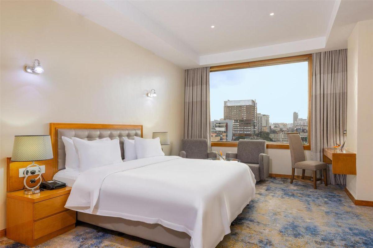A modern hotel room with a large bed in white linens, a patterned blue and gold carpet, and a large picture window overlooking a city skyline.