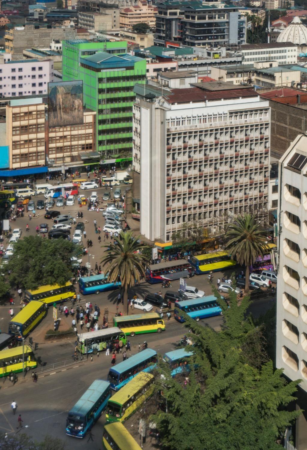 An aerial view of a busy city street intersection filled with numerous bright yellow and blue buses, surrounded by multi-story commercial buildings and scattered green trees.