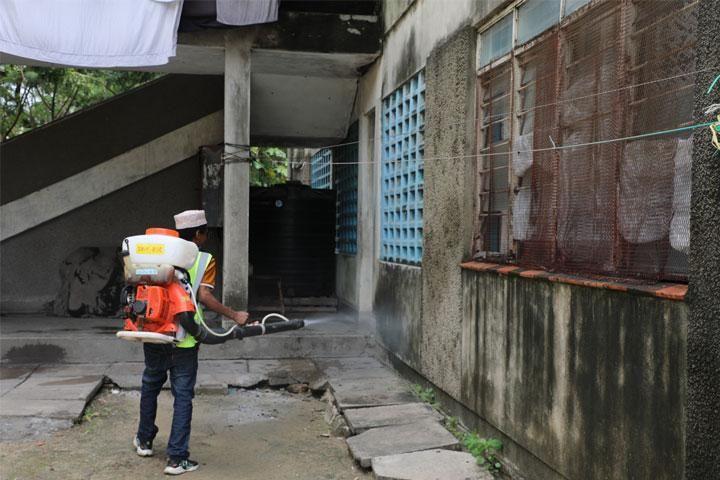 A worker wearing a safety vest and carrying a motorized backpack sprayer fumigates the exterior wall of a concrete building.