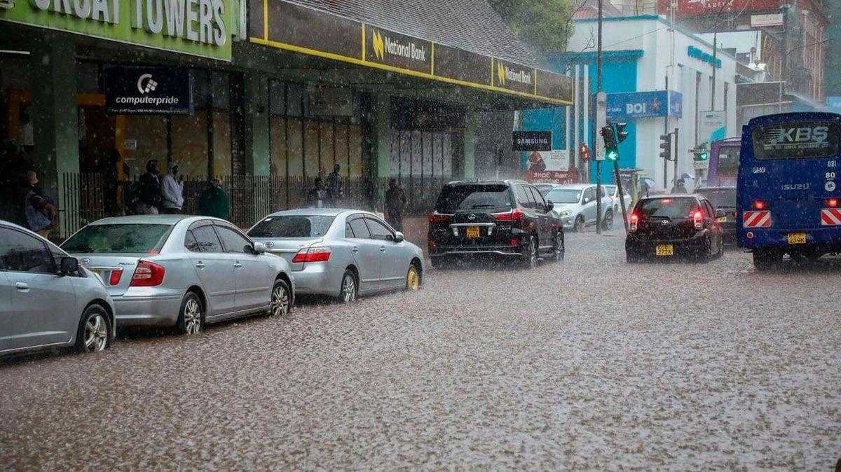 Cars and a blue bus drive through a heavily flooded city street during a torrential downpour next to commercial buildings.