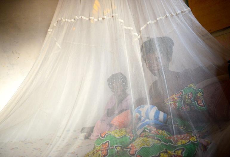 A mother sits with her young children on a bed underneath a large, white mosquito net.