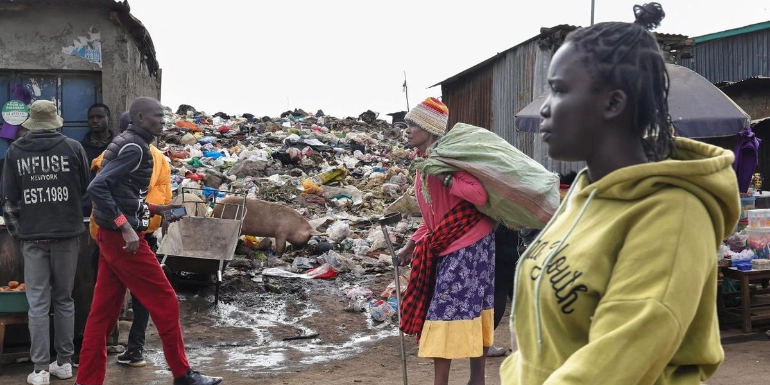 People walking along a muddy path next to a massive, sprawling pile of trash in an urban area.
