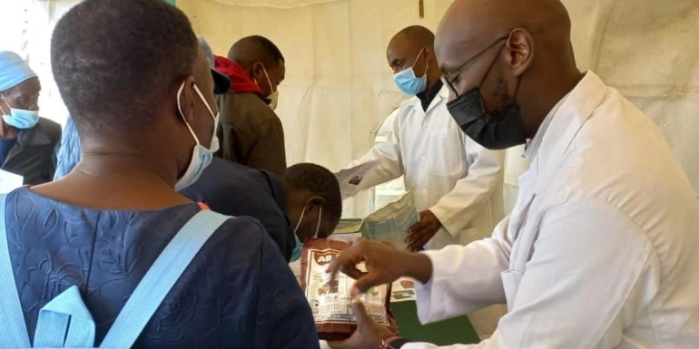 Healthcare workers in white lab coats and face masks hand out supplies to people under a large tent.