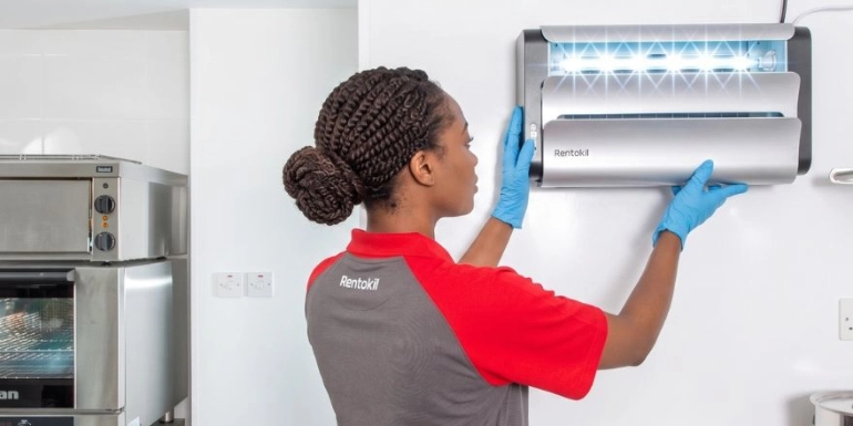 An African female Rentokil technician wearing a grey and red uniform and blue gloves installs a Lumnia fly control unit on a commercial kitchen wall.